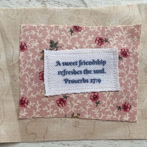 Puede incluir: Un cuadrado de tela con un fondo beige claro, un centro con estampado floral rosa y una etiqueta blanca. La etiqueta dice "A sweet friendship refreshes the soul. Proverbios 27:9" en texto azul. El estampado floral incluye flores rojas.