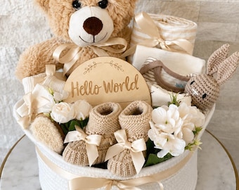 Può includere: Un cesto intrecciato bianco contenente un orsacchiotto color marrone chiaro, una targa in legno con la scritta Hello World, un paio di scarpine in maglia con fiocchi in raso, un sonaglio a forma di coniglio all'uncinetto e fiori artificiali.