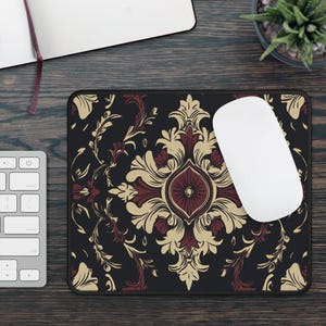 May include: A black mousepad with a decorative floral pattern in burgundy and gold. A white wireless mouse rests on the pad, next to a keyboard and a notebook. The mousepad has a rectangular shape.
