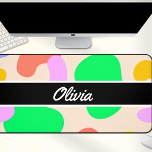 May include: A rectangular desk mat with a beige background and colorful abstract shapes in green, pink, orange, and yellow. A black stripe across the center displays the name "Olivia" in white script. A computer, keyboard, and mouse are visible.