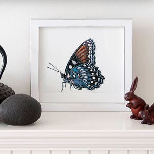 May include: A framed butterfly art print with a white frame, featuring a detailed illustration of a butterfly with blue, brown, and white markings. Beside the frame are a dark brown teapot, a dark gray stone, and two small brown rabbit figurines.
