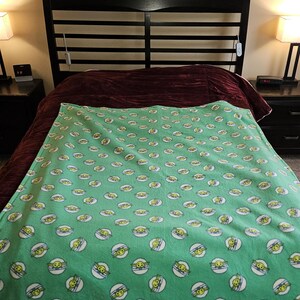 K&ouml;nnte beinhalten: Eine gr&uuml;ne Fleecedecke mit einem sich wiederholenden Muster eines Cartoon-Gesichts in einem wei&szlig;en Kreis mit blauem Umriss. Die Decke liegt auf einem Bett mit einem dunkelbraunen Kopfteil und einer weinroten Tagesdecke.