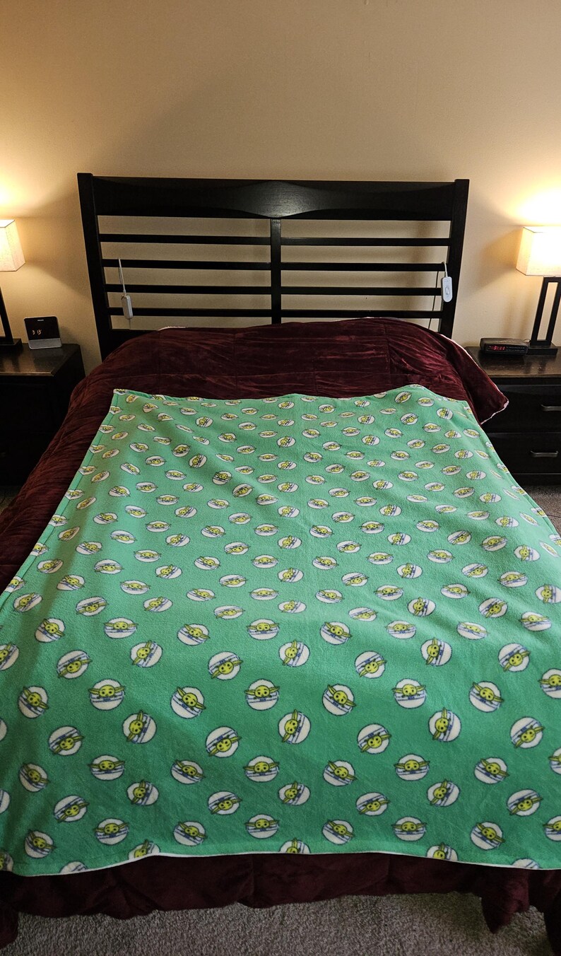 K&ouml;nnte beinhalten: Eine gr&uuml;ne Fleecedecke mit einem sich wiederholenden Muster eines Cartoon-Gesichts in einem wei&szlig;en Kreis mit blauem Umriss. Die Decke liegt auf einem Bett mit einem dunkelbraunen Kopfteil und einer weinroten Tagesdecke.