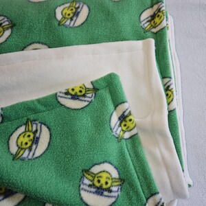 K&ouml;nnte beinhalten: Eine gr&uuml;ne Fleece-Decke mit einem sich wiederholenden Muster eines gelben Charakters mit gro&szlig;en Ohren in einem wei&szlig;en und blauen Oval. Die Decke ist gefaltet, um eine einfarbige wei&szlig;e Fleece-Schicht freizulegen. Die Decke ist weich und gem&uuml;tlich.