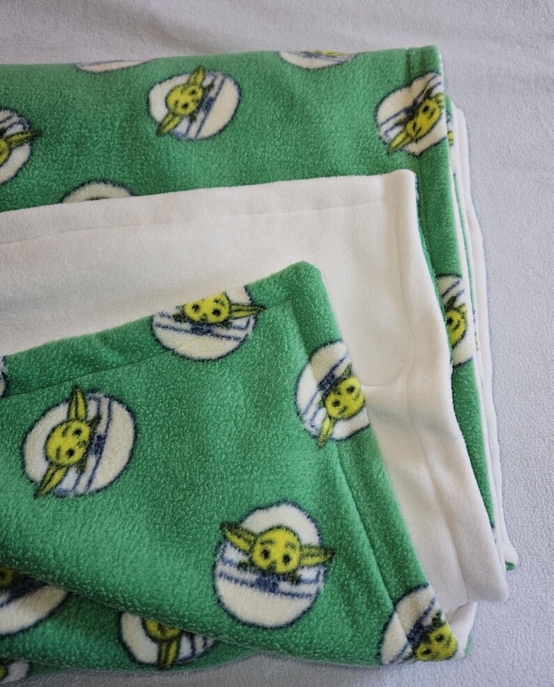 K&ouml;nnte beinhalten: Eine gr&uuml;ne Fleece-Decke mit einem sich wiederholenden Muster eines gelben Charakters mit gro&szlig;en Ohren in einem wei&szlig;en und blauen Oval. Die Decke ist gefaltet, um eine einfarbige wei&szlig;e Fleece-Schicht freizulegen. Die Decke ist weich und gem&uuml;tlich.