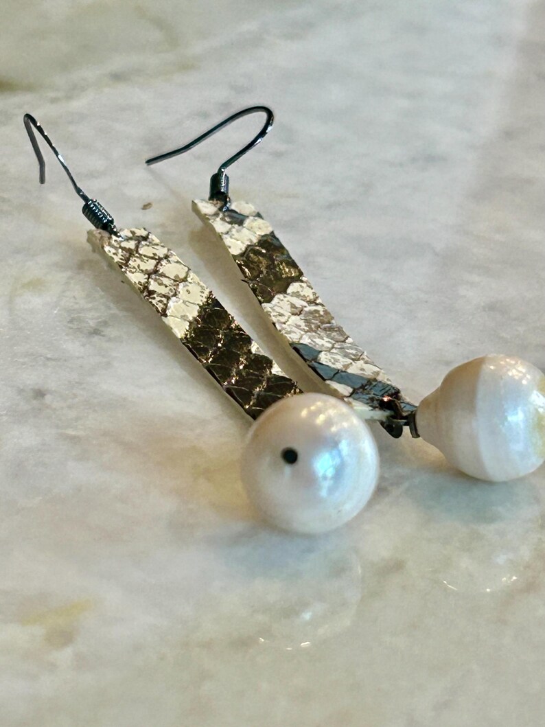 Może przedstawiać: Para kolczyk&oacute;w z wiszącymi elementami ze wzorem sk&oacute;ry węża i białą perłą. Kolczyki mają długą, prostokątną g&oacute;rę ze wzorem sk&oacute;ry węża z białą perłą na dole. Haczyki na uszy są czarne.