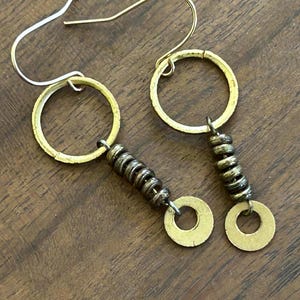 May include: A pair of handmade earrings with gold-coloured hoops and small, stacked, dark-coloured beads. The earrings have a unique, artistic design with a circular element and a hook closure. The earrings are displayed on a wooden surface.