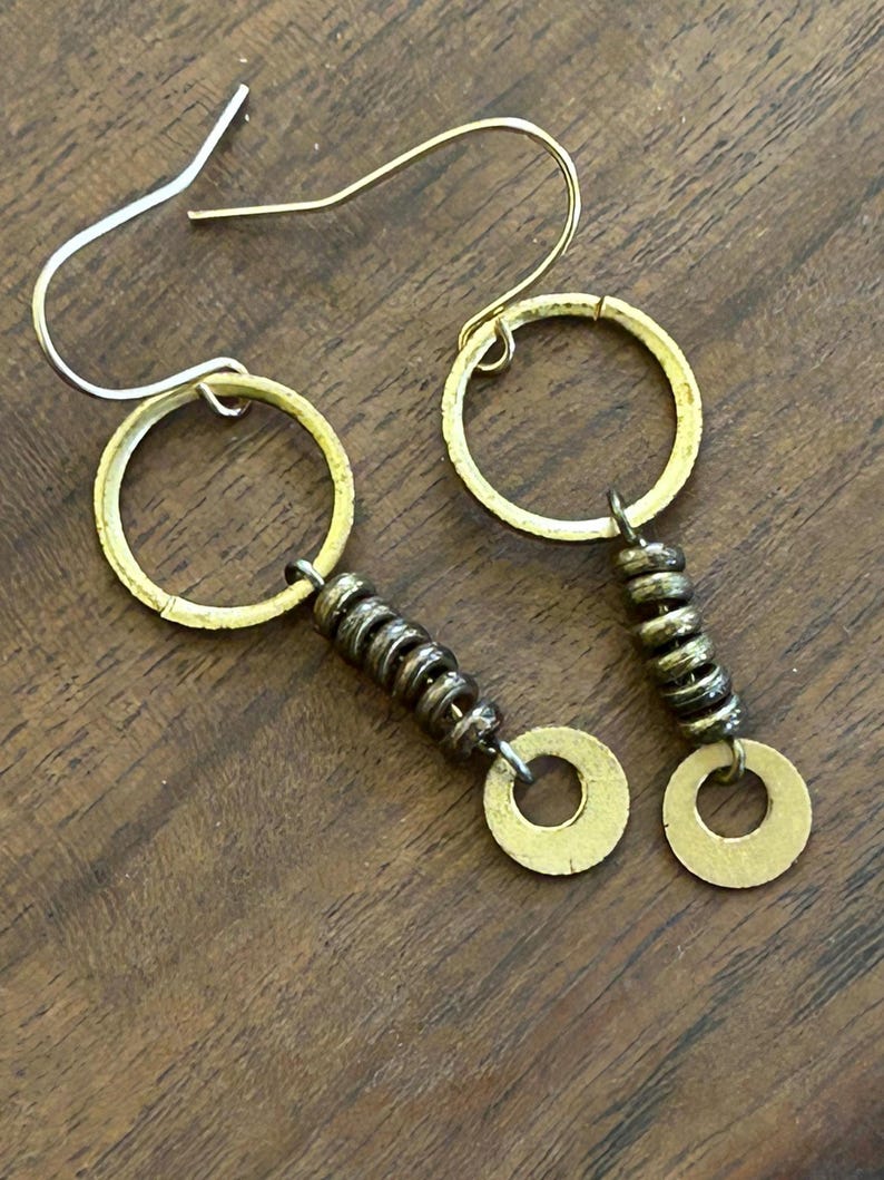 May include: A pair of handmade earrings with gold-coloured hoops and small, stacked, dark-coloured beads. The earrings have a unique, artistic design with a circular element and a hook closure. The earrings are displayed on a wooden surface.