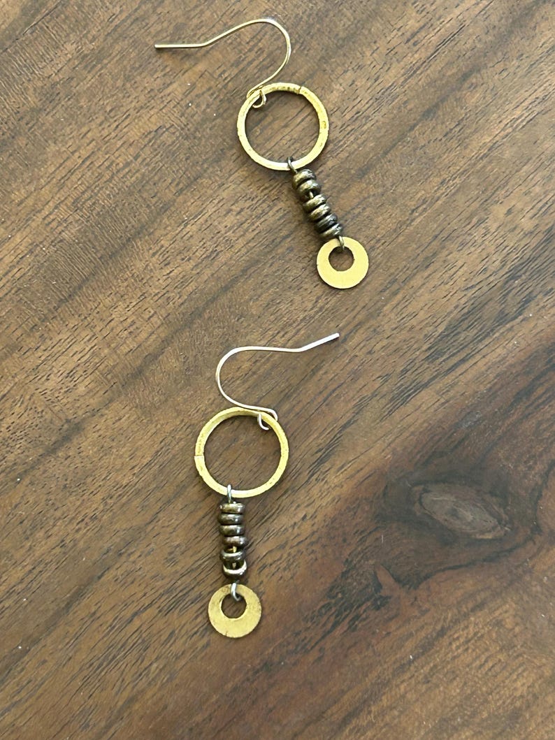 May include: A pair of earrings with a distinctive design. Each earring has a gold-coloured circular hoop, connected to a series of small, dark beads, and a small, flat, gold-coloured circle at the bottom. The earrings are displayed on a wooden surface.