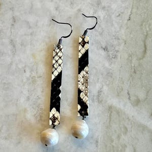 May include: A pair of dangle earrings with a snakeskin pattern in black and white. The earrings have a rectangular shape with a pearl-like bead at the bottom and black ear wires.
