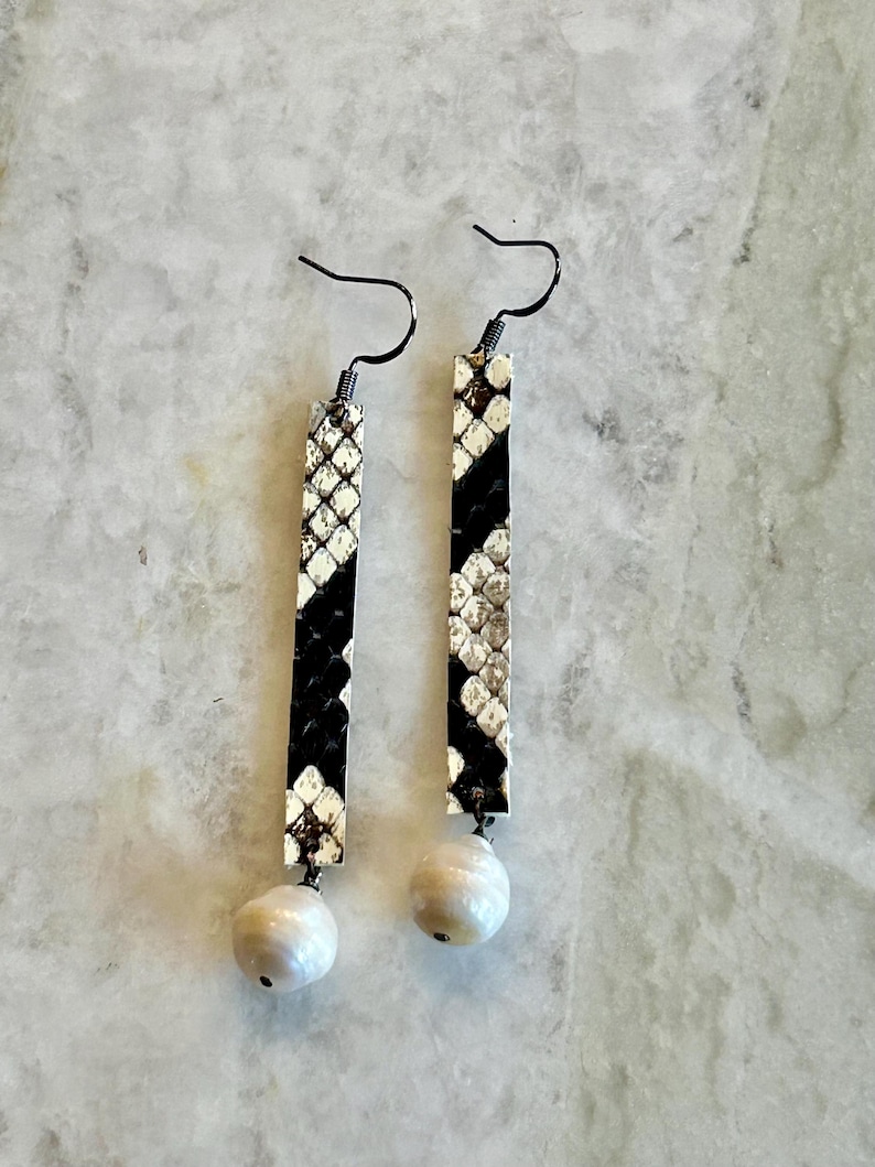 Może przedstawiać: Kolczyki wiszące z wzorem sk&oacute;ry węża w kolorze czarno-białym. Prostokątne kolczyki mają perłową kulkę na dole i czarne haczyki.