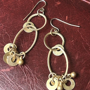 May include: A pair of gold-toned earrings with a unique design. Each earring features an elongated oval shape, connected to a smaller circle and a fish hook ear wire. Small, round gold beads and flat, circular charms dangle from the bottom of the oval.