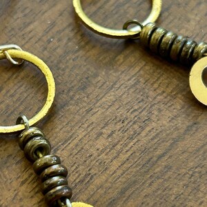 May include: Close-up of gold earrings with a circular design. The earrings feature small, dark-coloured beads and a larger, flat, circular element. The earrings are set against a wooden background.