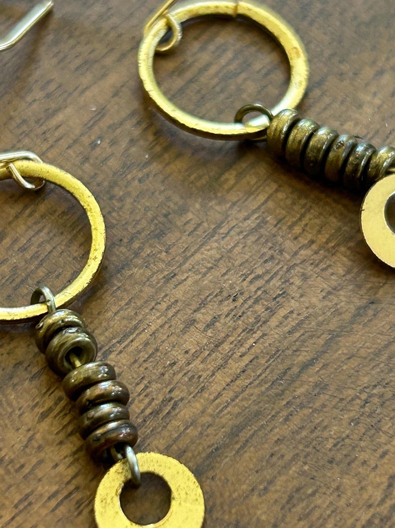 May include: Close-up of gold earrings with a circular design. The earrings feature small, dark-coloured beads and a larger, flat, circular element. The earrings are set against a wooden background.