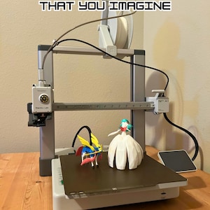 May include: A white 3D printer with a spool of filament, a black platform, and two 3D printed figurines. The figurines are a blue and yellow creature and a white dress with a green head. The printer has the text "YOU CAN CREATE ANYTHING THAT YOU IMAGINE" above it.
