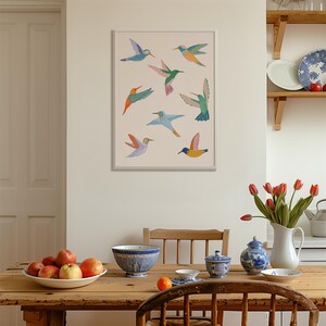 Op de afbeelding: Een ingelijste wandkunstprint met acht gestileerde kolibries in diverse kleuren, waaronder blauw, groen, oranje, roze en geel, gerangschikt op een lichtbeige achtergrond. De vogels zijn afgebeeld in vlucht met gespreide vleugels.