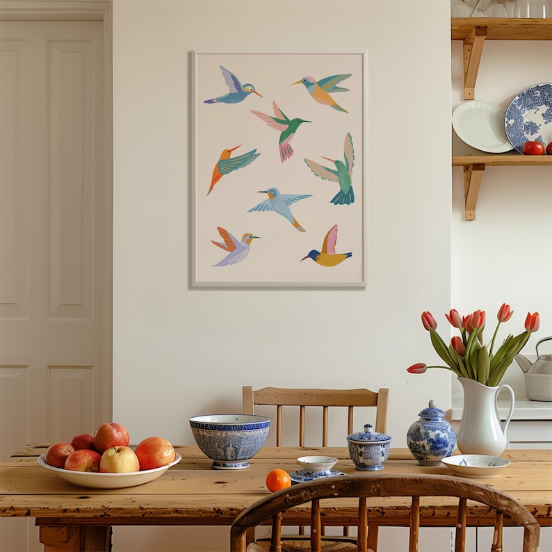 Op de afbeelding: Een ingelijste wandkunstprint met acht gestileerde kolibries in diverse kleuren, waaronder blauw, groen, oranje, roze en geel, gerangschikt op een lichtbeige achtergrond. De vogels zijn afgebeeld in vlucht met gespreide vleugels.