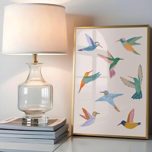 Op de afbeelding: Een tafellamp met een heldere glazen voet en een witte stoffen kap staat naast een ingelijste print. De print toont zeven kleurrijke kolibries in verschillende vliegposities, uitgevoerd in blauw-, groen-, oranje- en rozetinten op een witte achtergrond.