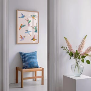 Op de afbeelding: Een ingelijste wandkunstprint met acht gestileerde kolibries in diverse kleuren zoals blauw, oranje, groen en roze op een lichte achtergrond. Daaronder ligt een blauw-wit geblokt sierkussen op een houten krukje.