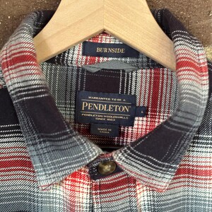 May include: A close-up of a plaid shirt in red, grey, and black, hanging on a wooden hanger. The shirt features the label "PENDLETON WOOLEN MILLS SINCE 1863". The collar has a "BURNISDE" label.