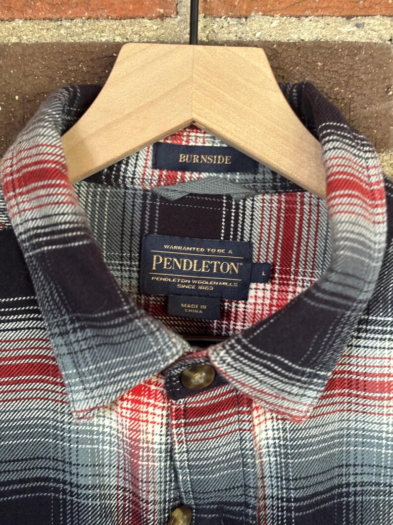 May include: A close-up of a plaid shirt in red, grey, and black, hanging on a wooden hanger. The shirt features the label "PENDLETON WOOLEN MILLS SINCE 1863". The collar has a "BURNISDE" label.