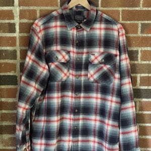 以下が含まれることがあります： 赤、白、ネイビーブルーのチェック柄長袖シャツ。シャツには胸ポケットが2つあり、木製のハンガーに吊るされています。柄は伝統的なチェック柄です。