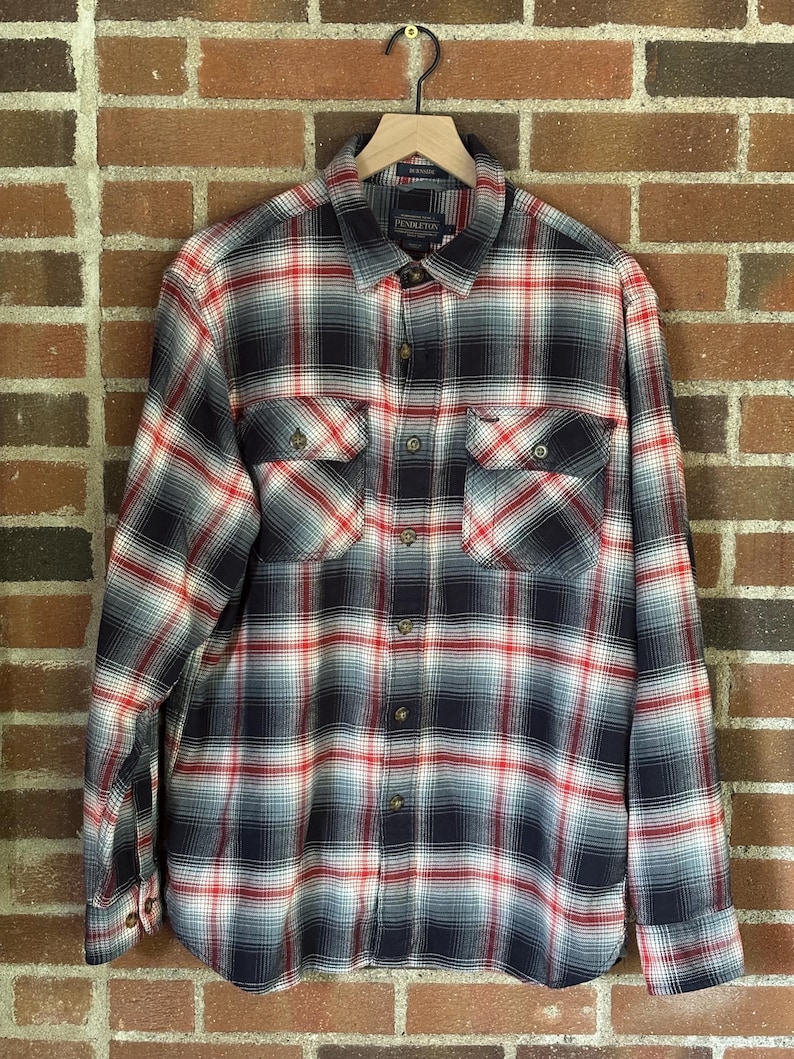 May include: A long-sleeved, button-down plaid shirt in red, white, and navy blue. The shirt has two chest pockets and is suspended from a wooden hanger. The shirt's pattern is a traditional check design.