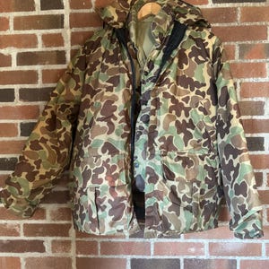 Puede incluir: Chaqueta de camuflaje con capucha, colgada contra una pared de ladrillos. La chaqueta presenta un patrón de colores marrón, verde y beige. Tiene cierre de cremallera y varios bolsillos. Diseñada para actividades al aire libre.
