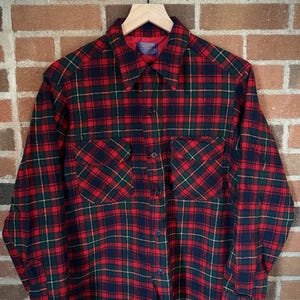 Può includere: Camicia di flanella a quadri rossi, verdi e neri con chiusura a bottoni e due tasche sul petto. La camicia ha un'etichetta che recita "Pendleton".