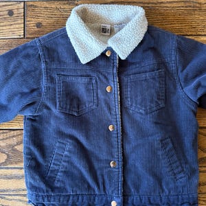 May include: Navy blue corduroy jacket with a cream-colored, fuzzy collar. The jacket has four front pockets, gold-colored buttons, and a soft, textured lining. The jacket is a children's size 4-6.