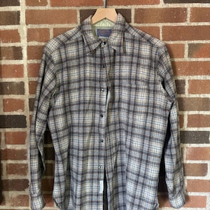 May include: A button-down flannel shirt with a plaid pattern in shades of brown, blue, and beige. The long-sleeved shirt is hanging on a wooden hanger against a brick wall background.