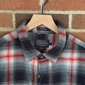 May include: A checked shirt in red, white, and navy blue, hanging on a wooden hanger. The shirt features a collared neckline and buttoned cuffs. The brand name "Pendleton" is visible on the label.