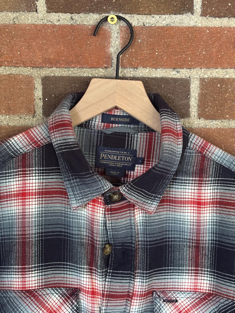 May include: A checked shirt in red, white, and navy blue, hanging on a wooden hanger. The shirt features a collared neckline and buttoned cuffs. The brand name "Pendleton" is visible on the label.