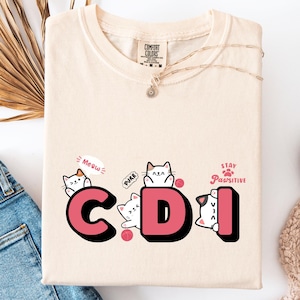 May include: Cream-colored t-shirt with the letters "CDI" in pink, featuring cartoon cat illustrations. The shirt also includes the phrases "Meow," "Purr," and "Stay Pawsitive." The shirt is folded on a white surface next to a pair of blue jeans.