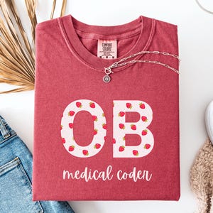 May include: A red t-shirt with the letters "OB" filled with strawberry patterns, and the words "medical coder" in white. The shirt is folded and placed on a white surface with a pair of blue jeans and a necklace.