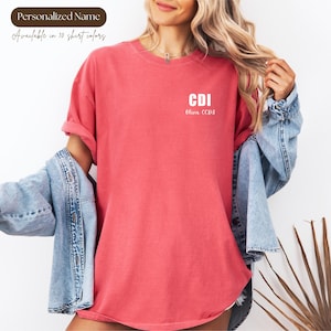 May include: Coral-colored t-shirt with the text "CDI Olivia CCPS" in white. The shirt has rolled-up sleeves and a relaxed fit. The image also shows the text "Personalized Name" and "Available in 10 shirt colors".