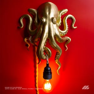 May include: A golden octopus wall sconce with a hanging lightbulb against a red background. The lightbulb is lit, and the fixture is attached to the wall with a rope. The text "MADE IN CALIFORNIA WITH CARE BY OUR SMALL FAMILY BUSINESS" is at the bottom.