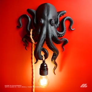 May include: Black octopus wall lamp with a glowing lightbulb hanging from its tentacles. The lamp is mounted on a red wall. The text "MADE IN CALIFORNIA WITH CARE BY OUR SMALL FAMILY BUSINESS" is at the bottom.