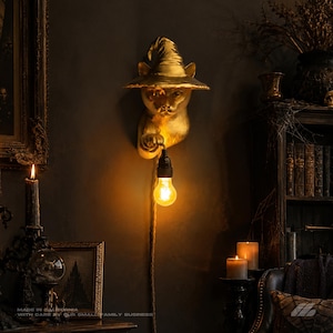 May include: A gold-colored wall lamp shaped like a cat wearing a pointed witch hat. The cat figure is mounted to the wall and holds a black socket with a glowing, warm-toned Edison-style light bulb suspended by a braided cord hanging downward.