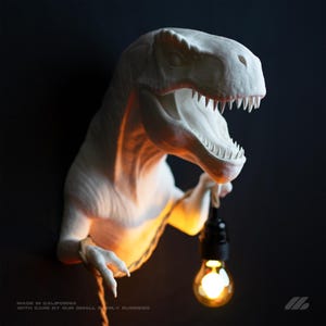 May include: A white wall-mounted Tyrannosaurus Rex sculpture featuring a textured finish and an open mouth. The dinosaur holds a black light socket with a glowing light bulb suspended by a rope. Text at the bottom reads: MADE IN CALIFORNIA WITH CARE BY OUR SMALL FAMILY BUSINESS.