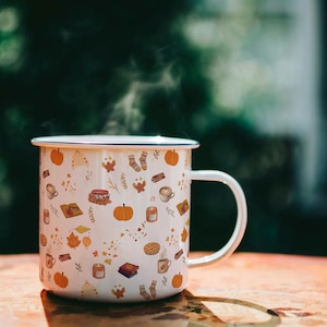 Puede incluir: Una taza de esmalte blanco con asa, que presenta un patrón repetitivo de calabazas naranjas, libros marrones, hojas de otoño amarillas, velas y calcetines. El vapor sale de la parte superior de la taza, que tiene un borde oscuro fino.