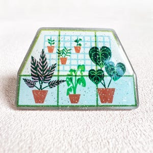 May include: A rectangular pin featuring a greenhouse design with various potted plants. The pin has a light blue background with a grid pattern and glitter accents. The plants are in brown pots, and the pin has a clear, glittery border.