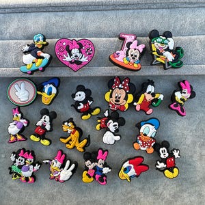 May include: A selection of colourful cartoon character shoe charms featuring Mickey Mouse, Minnie Mouse, Donald Duck, and other Disney characters. The charms are in various poses and designs, with bright colours and detailed features.