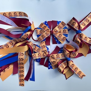 May include: A collection of hair bows in different designs and colours. Some bows are made with ribbons printed with a lion graphic and the text "LIONS". The bows are in shades of gold, burgundy, blue, and white. Some bows have long, flowing ribbons.