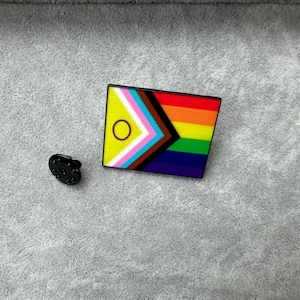 May include: A black enamel pin featuring a rainbow flag design. The flag has a yellow triangle with a white circle in the centre, followed by pink, blue, white, brown, black, and the rainbow colours.