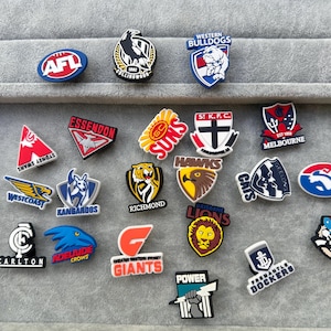 May include: A selection of AFL team logo charms in various colours. The charms display team logos in red, blue, yellow, and black. These are likely decorative accessories for shoes or other items.