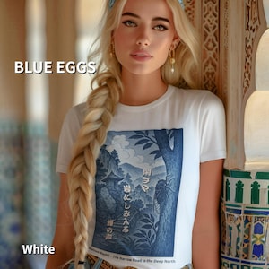 Puede incluir: Camiseta blanca con estampado gráfico azul y blanco con un árbol, montañas y texto japonés. También se ven los textos "BLUE EGGS" y "White". La persona lleva una camiseta con pelo largo y rubio.