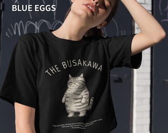 Puede incluir: Camiseta negra con una ilustración de un gato gruñón y el texto "THE BUSAKAWA". El gato es gris y blanco con una expresión de mal humor. La camiseta tiene cuello redondo y una cita debajo.