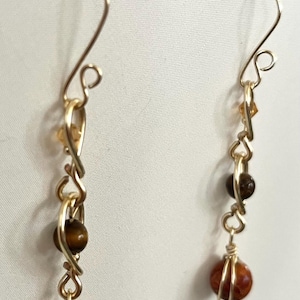 Peut inclure: Une paire de boucles d'oreilles artisanales avec du fil doré et des perles en pierre naturelle. Les boucles d'oreilles présentent un design à crochet, avec une série de petites perles et une pierre plus grande, ronde, brun-rougeâtre en bas.