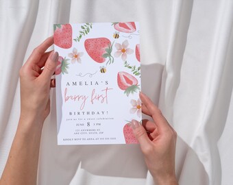Puede incluir: Una tarjeta de invitación blanca con un diseño de fresa rosa y verde. El texto en la tarjeta dice "Amelia's Berry First Birthday!" con la fecha y la hora del evento.